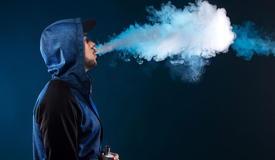 Mann in einer blauen Kapuzenjacke, der eine große Dampfwolke ausatmet, während er eine Vape in der Hand hält, vor einem dunkelblauen Hintergrund