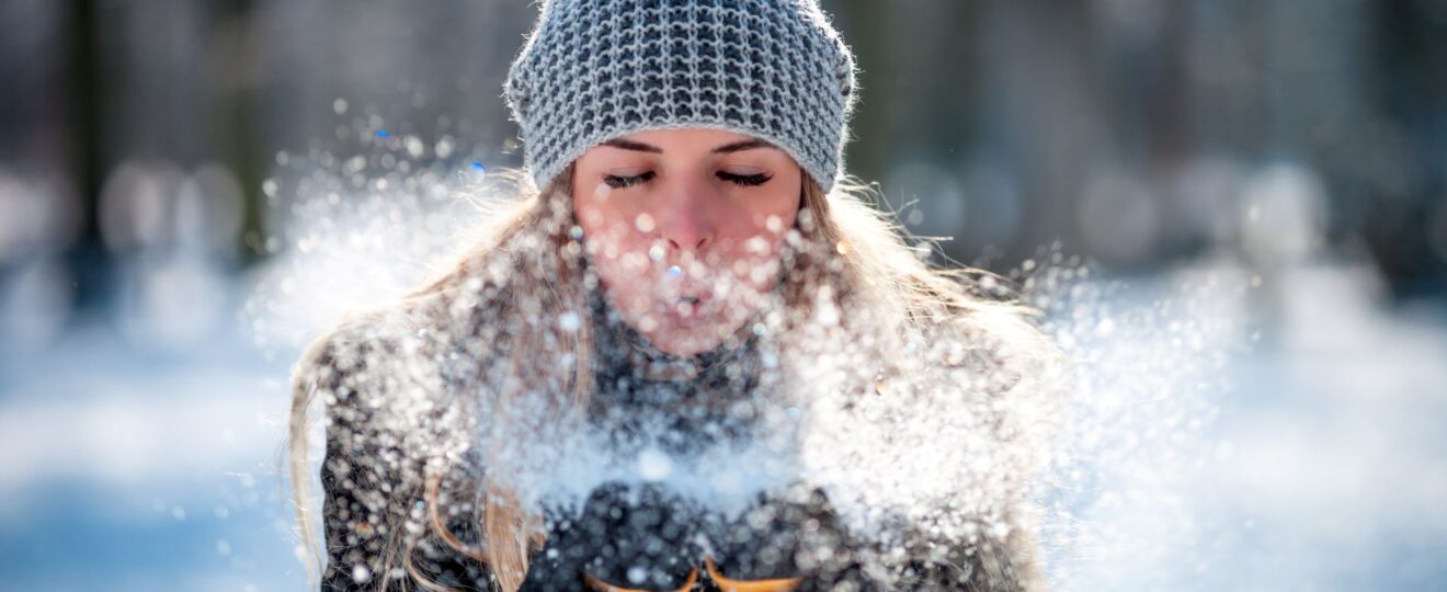 Frau mit Alpaka Mütze bläst Schnee in winterlicher Kulisse.