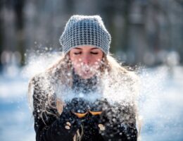 Frau mit Alpaka Mütze bläst Schnee in winterlicher Kulisse.