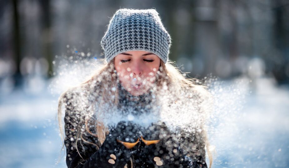 Frau mit Alpaka Mütze bläst Schnee in winterlicher Kulisse.