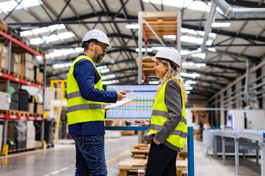 Mitarbeiterin und Manager inspizieren Produktionsprozesse mit automatisierten Maschinen in einer modernen Lagerhalle.