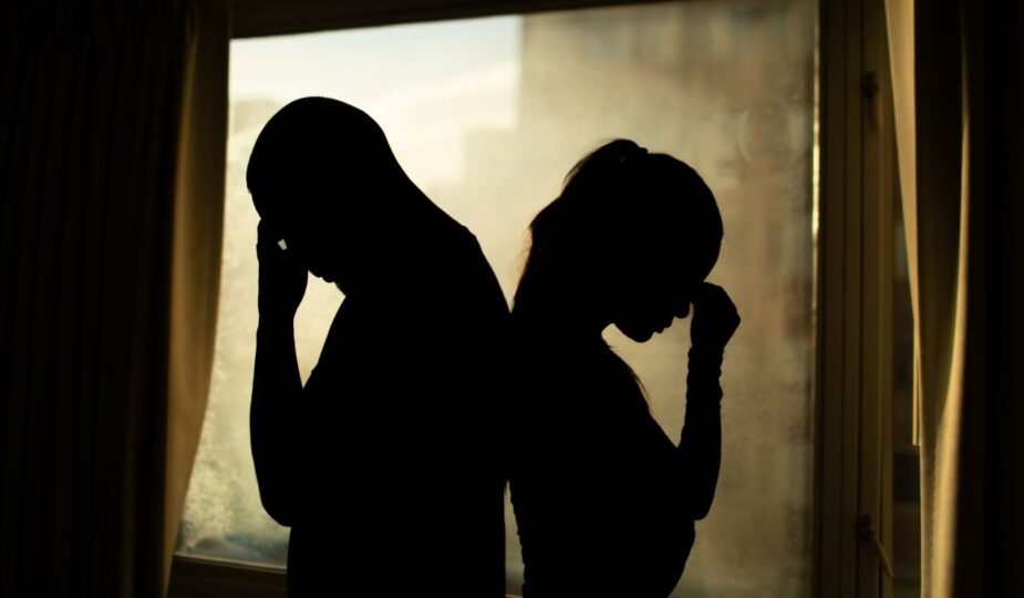 Mann und Frau stehen rueckwaerts vor dem Fenster und halten ihre Koepfe – Symbolbild fuer emotionale Belastung und versteckte Scheidung Kosten